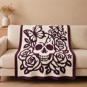 May include: A rectangular crochet blanket featuring a dark purple and cream color scheme. The design displays a central human skull surrounded by blooming roses and butterflies, finished with a solid dark purple border along the edges.