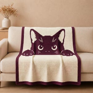 Puede incluir: Una manta de ganchillo rectangular con un gráfico de gato de color morado oscuro centrado sobre un fondo de color crema. El diseño del gato muestra una cabeza con orejas puntiagudas y dos patas asomando por el borde. La manta tiene un borde morado.
