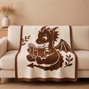 Puede incluir: Una manta de ganchillo color crema con un gráfico pixelado marrón oscuro de un pequeño dragón leyendo un libro abierto. La manta tiene un borde sólido de color marrón oscuro e incluye pequeños motivos decorativos de hojas a ambos lados.