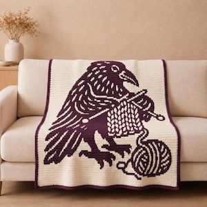 May include: A rectangular crochet blanket featuring a large, dark purple graphic of a raven knitting with two needles. The raven is depicted with a ball of yarn at its feet. The blanket has a cream-colored background and a dark purple border.