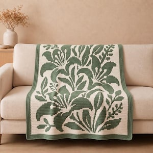 May include: A rectangular crochet blanket featuring a two-tone botanical pattern of large sage green lily flowers and leaves set against a cream-colored background. The throw has a solid sage green border framing the intricate floral design.