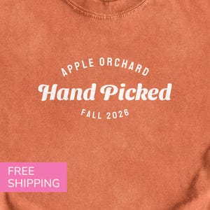 May include: A burnt orange crew neck t-shirt featuring white screen-printed text that reads APPLE ORCHARD Hand Picked FALL 2026. The fabric has a slightly weathered, vintage-inspired texture with a consistent solid color throughout the garment.