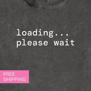 May include: A charcoal gray t-shirt featuring the white text "loading... please wait" printed in a typewriter-style font across the chest. The fabric has a slightly faded, textured appearance with a ribbed crew neckline.
