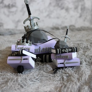 May include: A collection of miniature book ornaments in light purple and black, each featuring the word Fantasy in raised lettering, silver-colored swords, and crowns. Some books are topped with gray stone-like shapes and silver swords.