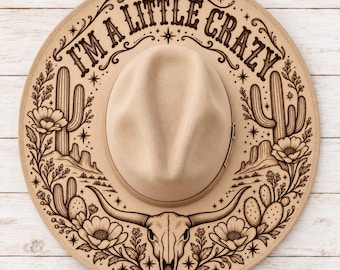May include: A tan felt hat featuring a dark brown laser-engraved design of a longhorn skull, cacti, and desert flowers. The text "I'M A LITTLE CRAZY" is arched across the top, surrounded by small star accents and decorative flourishes.