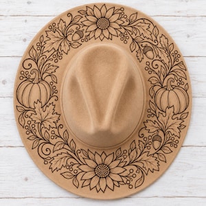 Op de afbeelding: Een beige vilten hoed met een donkerbruin, lasergegraveerd ontwerp op de rand. Het patroon bestaat uit een cirkelvormig kransmotief van zonnebloemen, pompoenen, esdoornbladeren, eikels en kleine bessen rondom de centrale kroon.