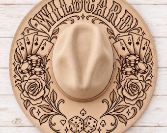 May include: A tan wide-brimmed hat featuring a dark brown laser-engraved design. The brim displays the text WILDCARD, playing cards, stacks of poker chips, roses, dice, and card suit symbols arranged in a symmetrical circular pattern.