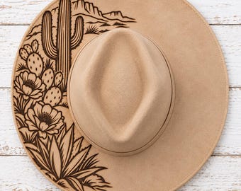 May include: A tan wide-brimmed hat featuring a dark brown laser-engraved desert scene on the left side of the brim. The design includes a tall saguaro cactus, prickly pear cacti with blooming flowers, agave plants, and distant mountain silhouettes.