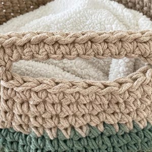 May include: A round, hand-crocheted storage basket featuring a colour-blocked design with a sage green base, a beige middle section, and a tan upper rim. The basket includes integrated side handles and contains a soft, white, textured fabric liner.