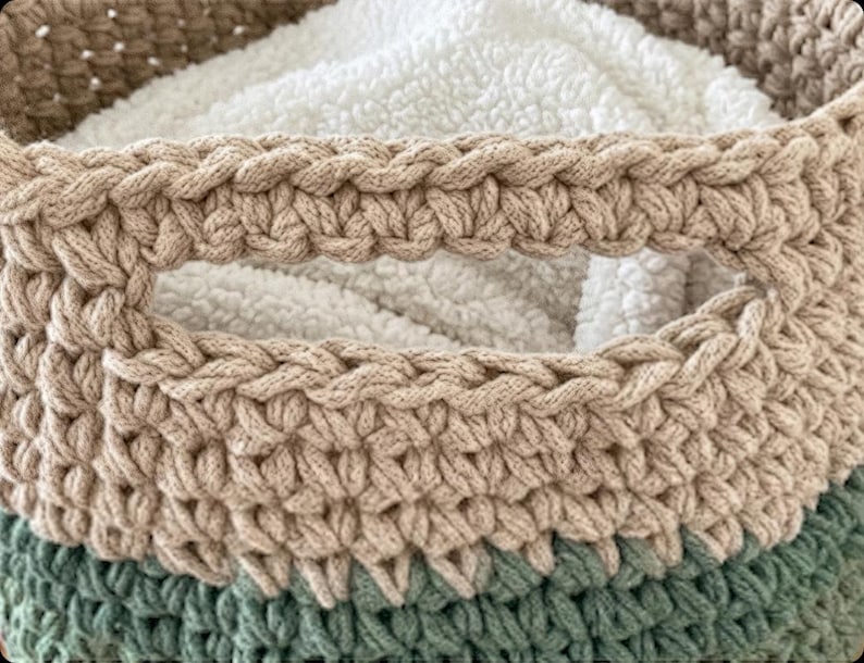 May include: A round, hand-crocheted storage basket featuring a colour-blocked design with a sage green base, a beige middle section, and a tan upper rim. The basket includes integrated side handles and contains a soft, white, textured fabric liner.