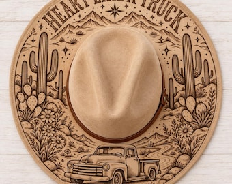 May include: A tan felt hat featuring a dark brown band and a laser-engraved desert scene on the brim. The design includes cacti, mountains, flowers, and a vintage pickup truck with the text HEART LIKE A TRUCK arched across the top.