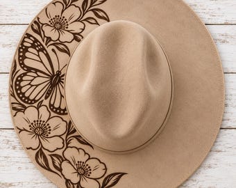 May include: A tan wide-brimmed hat featuring a dark brown laser-engraved design on the left side of the brim. The design consists of a large butterfly positioned between two blooming flowers with detailed petals and leaves extending outward.