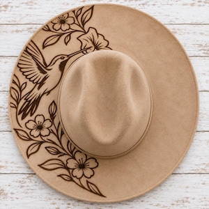 Puede incluir: Un sombrero de fieltro color canela con un diseño grabado a láser en color marrón oscuro en el ala. La ilustración muestra un colibrí en vuelo rodeado de elementos florales y hojas, extendiéndose desde la parte superior izquierda a la inferior derecha.