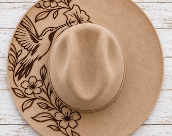 May include: A tan felt hat featuring a dark brown laser-engraved design on the brim. The artwork depicts a hummingbird in flight surrounded by floral elements and leaves, extending from the top left to the bottom right of the wide brim.