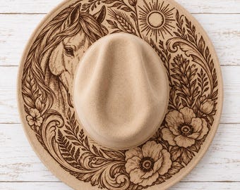 May include: A tan felt hat featuring a dark brown laser-engraved design on the brim. The artwork depicts a horse head surrounded by intricate floral patterns, wheat stalks, and a sunburst motif, all rendered in a detailed, illustrative style.