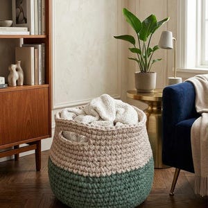 May include: A round, chunky knit storage basket featuring a two-tone design with a light beige upper section and a dark sage green lower section. The basket has two integrated side handles and is filled with a soft, white, textured fabric throw.