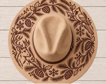 May include: A tan felt hat featuring a dark brown laser-engraved design on the brim, including a deer, a cardinal, pine branches, pinecones, and snowflakes. A dark brown band encircles the crown. Text at the bottom reads: SVG • PNG • Hand Burning PDFs.