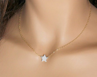 Puede incluir: Un delicado collar dorado con un pequeño colgante de estrella plateada. La estrella está centrada en una fina cadena, creando un diseño minimalista y elegante. El collar se muestra sobre un fondo color piel.