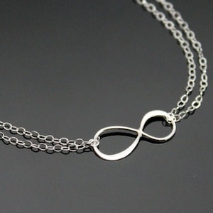 Könnte beinhalten: Halskette mit silbernem Unendlichkeitssymbol. Die Halskette hat ein Doppelketten-Design mit einem zarten Anhänger mit Unendlichkeitssymbol. Der Schmuck wird vor einem dunklen Hintergrund präsentiert.