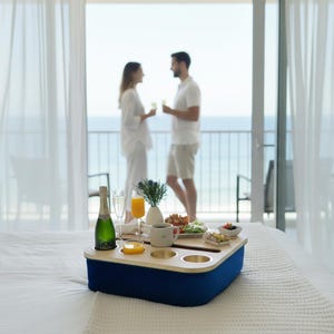 May include: A blue fabric bed tray with a light wood top featuring circular cutouts for a bottle of champagne, two glasses, a mug, and small bowls of food. The tray sits on a white bedspread in a room with a balcony view of the ocean.