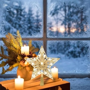 Op de afbeelding: Een vijfpuntige ster als piek voor in de kerstboom, voorzien van een helder, gefacetteerd kristalachtig ontwerp met geïntegreerde warmwitte ledverlichting. De ster staat op een houten ondergrond naast een dennentak en drie brandende kaarsen.