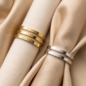 May include: Three gold-toned metal rings engraved with the names SOPHIE, LIAM, and MASON, and two silver-toned metal rings engraved with the names ELLA and NOAH. Each ring features a smooth, flat band design with clear, capitalized lettering.