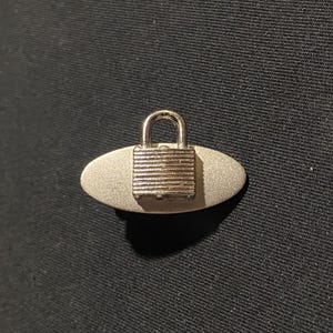 May include: A silver-toned metal button featuring a horizontal oval base with a textured surface and a small, raised padlock design centered on the front. The padlock has a classic shackle top and a rectangular body with horizontal grooves.
