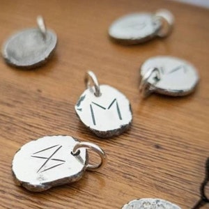 May include: A collection of silver charms with various engraved designs, including a letter M, a rune symbol, and a simple circle. The charms are displayed on a wooden surface.