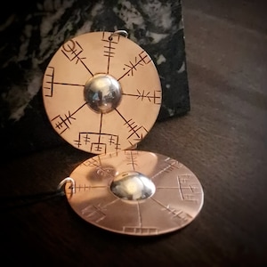 May include: Two round pendants made of copper with a silver centre. Each pendant features a circular design with a compass-like pattern and runes.