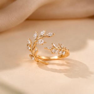 Puede incluir: Un anillo abierto de tono dorado que presenta dos ramas simétricas con piedras transparentes de corte marquesa engastadas en cada hoja. La banda está hecha de metal pulido y el diseño crea un efecto envolvente con un espacio abierto.
