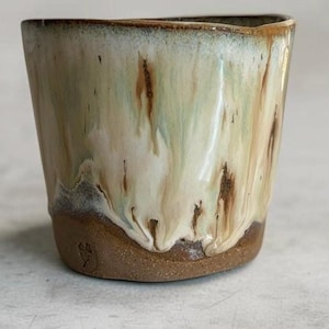 May include: A ceramic cup featuring a smooth, glossy glaze in shades of cream, pale green, and brown that drips down towards the base. The bottom portion of the cup is made of unglazed, textured brown clay with a small stamped maker's mark.
