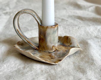 May include: A ceramic candle holder with a glossy, mottled glaze in shades of brown, cream, and grey. The piece features a wide, wavy-edged base, a central cylindrical socket, and a curved handle, holding a single white taper candle.