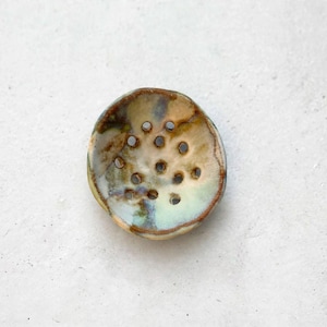 May include: A round, shallow ceramic soap dish featuring a mottled glaze in shades of tan, brown, and pale blue. The surface is punctuated by a series of small, evenly spaced circular drainage holes arranged in a cluster across the centre.