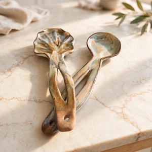 May include: Two handmade ceramic spoons with a glossy, marbled glaze in shades of brown, tan, and muted blue. Each spoon features a long, slender handle with a small circular hole at the end and a wide, shallow bowl with a scalloped edge design.