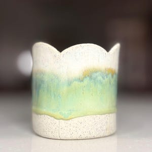 May include: A ceramic cup with a scalloped rim featuring a speckled off-white base and a smooth, flowing glaze transition from light green to a soft yellow-gold at the top edge. The cylindrical vessel has a textured, matte lower section.