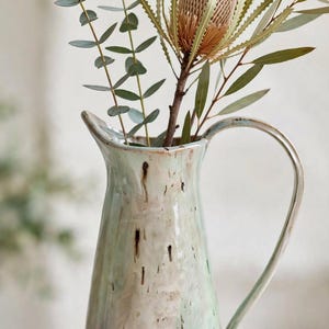 May include: A ceramic pitcher with a tapered body, a curved handle, and a spout. The glaze features a blend of pale green, white, and brown vertical streaks. The base is unglazed and textured, with the stamped text HM 24 visible near the bottom.