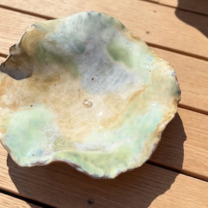 May include: A shallow, hand-formed ceramic bowl with an irregular, wavy rim. The surface features a glossy glaze in blended shades of pale mint green, soft yellow, and creamy white, creating a marbled effect across the interior basin.