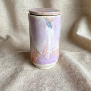 May include: A cylindrical ceramic canister with a matching lid, featuring a glossy glaze in shades of lavender, soft pink, and cream. The colours are applied in a fluid, dripping pattern that runs vertically down the exterior surface of the vessel.
