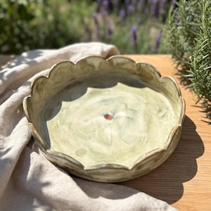 May include: A round ceramic dish with a scalloped edge and a light green, mottled glaze. The interior surface features a subtle, swirling texture with a small, centred brown circular mark, creating a flower-like appearance in the basin.