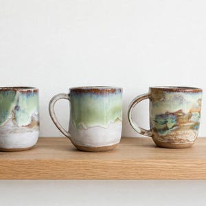 May include: Three ceramic mugs with a rounded shape and a handle on the side. Each mug features a reactive glaze finish with a colour palette of sage green, white, and earthy brown tones, showing a distinct drip pattern along the exterior surface.