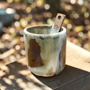 May include: A cylindrical ceramic cup with a reactive glaze featuring blended tones of cream, sage green, and deep brown. The cup has a smooth, rounded rim and a matching ceramic spoon with a circular hole at the top handle resting inside.