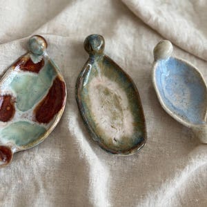 Può includere: Tre piccoli piatti in ceramica di forma ovale con finitura lucida. Il piatto a sinistra presenta un motivo maculato marrone e verde chiaro. Il piatto centrale ha un centro bianco con bordi verde scuro. Il piatto a destra è azzurro.