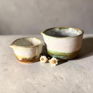 May include: Two ceramic pouring bowls with a rounded shape and a small spout on the side. The bowls feature a layered glaze finish with white, light green, and earthy brown tones. Two small white dried flowers are placed between the two bowls.