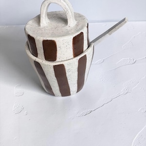 May include: A ceramic sugar bowl with a matching lid and a small spoon. The bowl and lid feature a speckled off-white glaze with vertical dark brown stripes. The lid has a prominent arched handle, and a spoon rests inside the bowl.