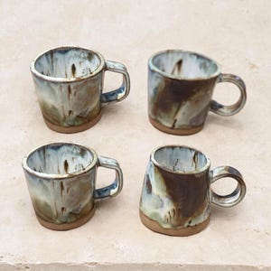 May include: A set of four ceramic mugs featuring a reactive glaze with swirling patterns of white, cream, and dark brown. Each mug has a sturdy handle and a smooth, unglazed base, showcasing a distinct, hand-thrown appearance with organic shapes.