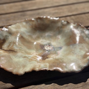 Puede incluir: Un cuenco de cerámica poco profundo, formado a mano, con un borde irregular y ondulado. La superficie presenta un esmalte brillante y moteado en tonos marrón tierra, verde apagado y crema, con un pequeño sello circular en el centro.