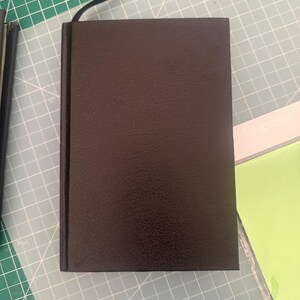 May include: A rectangular notebook with a dark brown, textured cover and a black ribbon bookmark attached to the spine. The notebook is positioned vertically, showcasing a solid, uniform color and a consistent, slightly grained surface texture.
