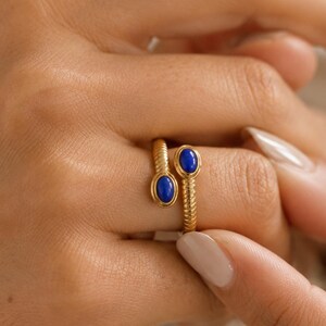 Puede incluir: Un anillo abierto de tono dorado con una banda de estilo cuerda trenzada. El anillo está decorado con dos pequeñas piedras ovaladas de color azul intenso, una en cada extremo del diseño abierto, creando un aspecto simétrico.