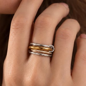Könnte beinhalten: Ein mehrreihiger Ring aus einer Kombination von polierten silber- und goldfarbenen Metallbändern. Das Design besteht aus mehreren gestapelten, ineinandergreifenden kreisförmigen Ringen, die ein geschichtetes Erscheinungsbild am Finger erzeugen und eine Mischung aus glatten metallischen Oberflächen und Texturen aufweisen.
