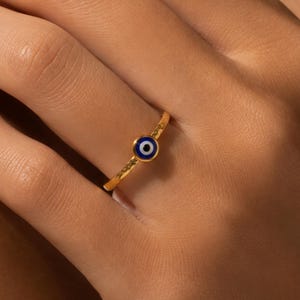 Puede incluir: Un anillo de banda fina de oro con un pequeño dije circular de ojo turco. El dije consiste en un centro azul oscuro rodeado por un anillo blanco y un borde dorado fino, colocado centralmente en la banda delgada de metal dorado pulido.